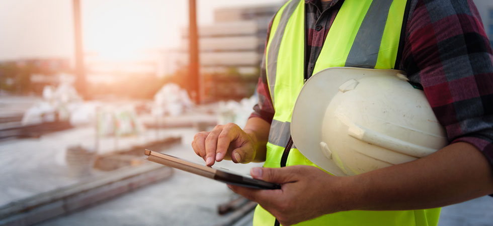 A construction worker wearing a bright safety vest has a hard hat tucked under their arm and is checking a tablet