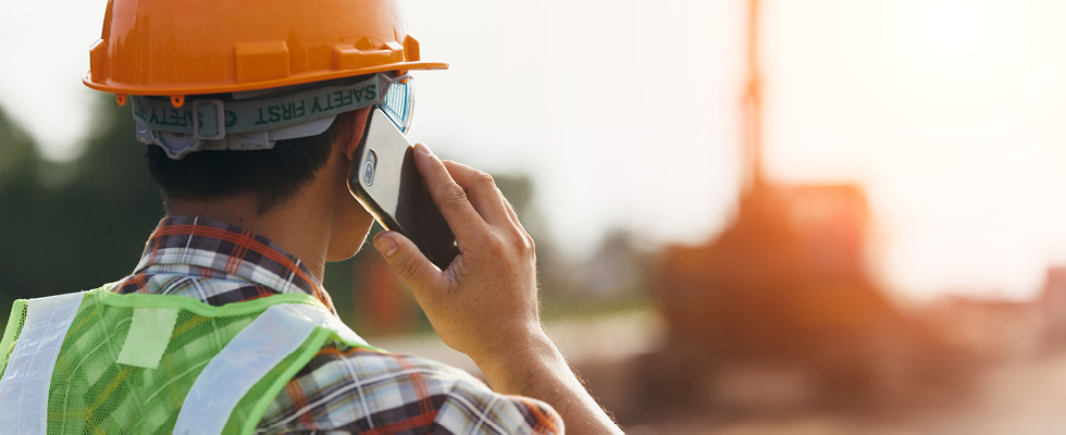 A person wearing an orange hardhat and a bright green safety vest, facing away from the camera, is holding a cellphone to their ear.