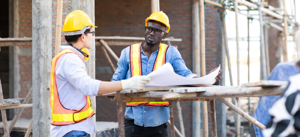 Two people wearing hard hats and bright safety vests talk over a large sheet over paper on a construction site