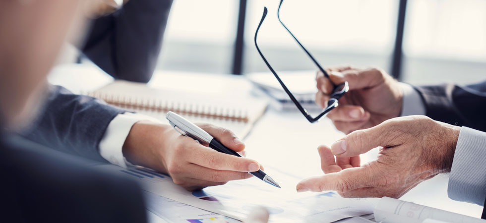 Two sets of hands point to graphs on a sheet of paper. One hand holds a pen. One hand holds a pair of glasses.