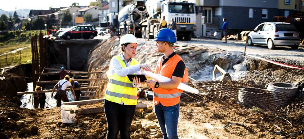 Two people in hard hats and bright safety vests are having a conversation while standing on a construction site