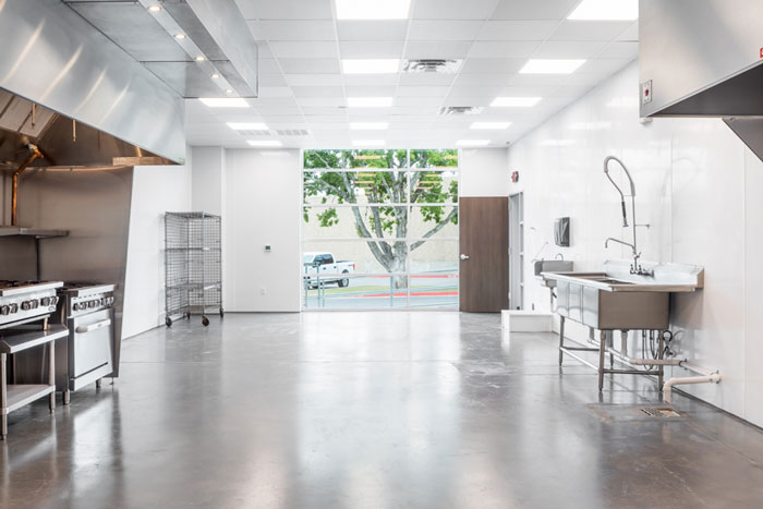 A bright room features an industrial work sink and two stovetops and ovens. A set of wire shelves sits in the back corner. A large window is at the back of the room near an open door. The floor is gray concrete and the walls are white.