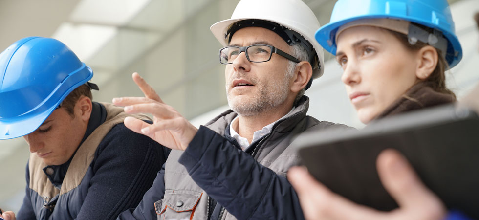 Two people wearing hardhats look forward. One of the people is wearing glasses and pointing at something out of frame. A third person, also wearing a hard hat, looks down.