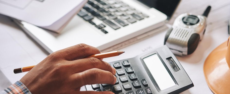 A person's hand is keying in numbers on a calculator. The hand is also holding a pencil. A portable radio and laptop can also be seen in the background.