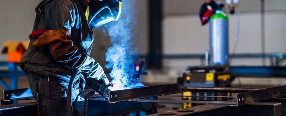 A worker welds something, illuminated in blue light