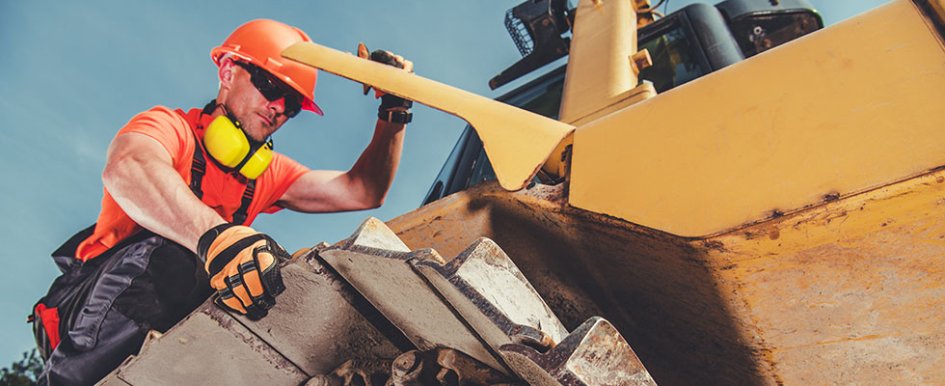 Worker in orange vest and hard hat with yellow headset, working on yellow piece of construction equipment