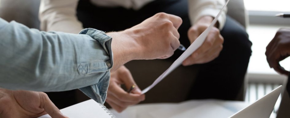 A hand uses a pen to point to a piece of paper that another set of hands is holding