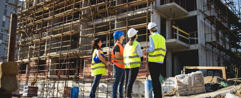 Four individuals look up at a building under construction. Three wear hardhats, but one does not.