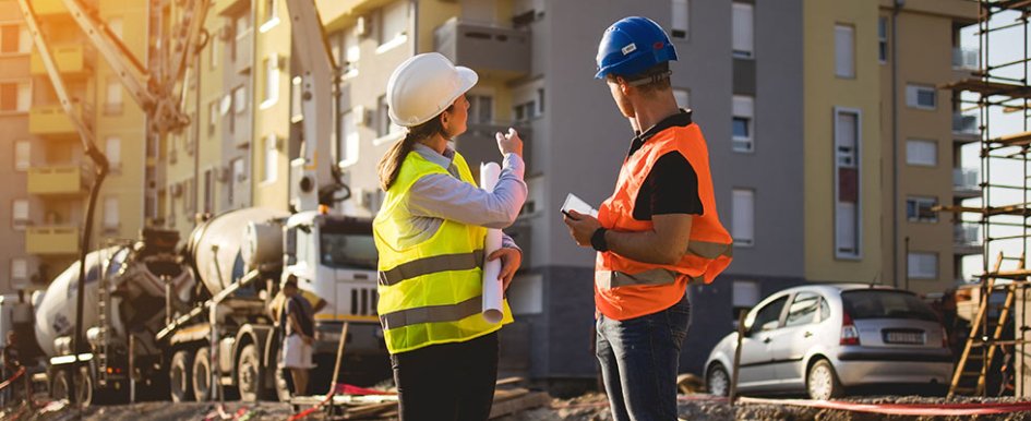 Two construction workers in hard hats and vests, one gesturing toward building, the other looking