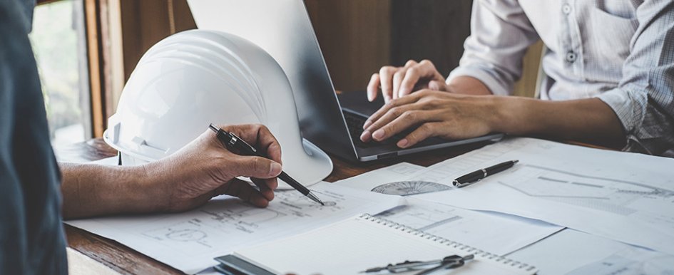 Two people working at desk with hard hat, plans on top of it