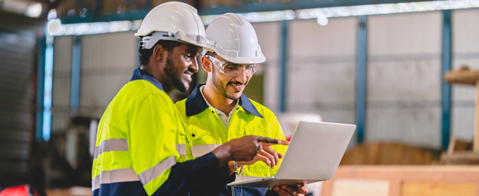 Two men in bright green work jackets, hard hats and safety gear looking at laptop