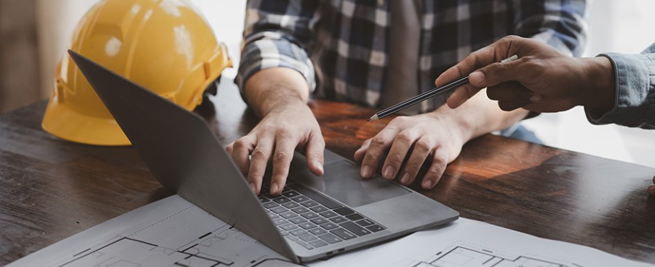 Person using laptop, with another person gesturing with a pencil. Yellow hard hat is beside them