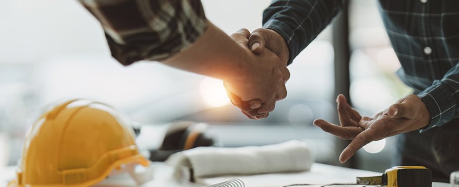 Two people shaking hands over a desk with hard hat and tape measure