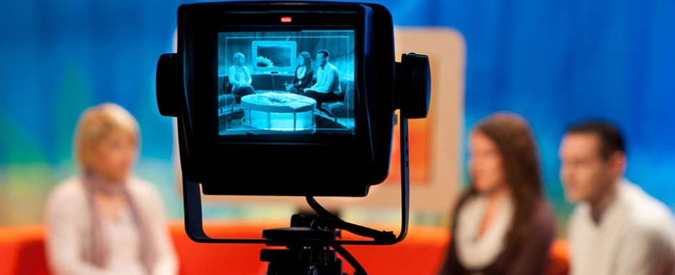A camera display shows a group of three people on a TV talk show