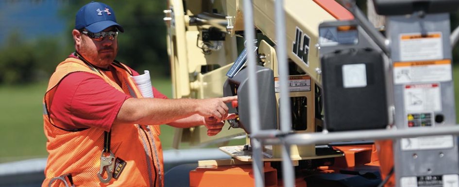 A construction worker in a bright safety vest and blue baseball hat presses a button on a piece of machinery