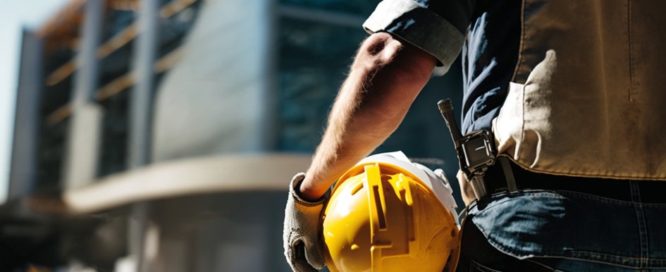 Person in gloves and vest holding yellow hard hat in front of building