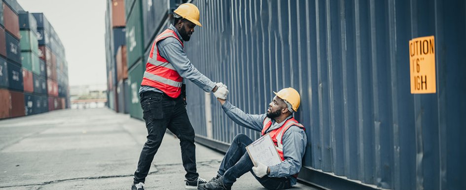 Two workers in vests and hard hats; one is being helped up by the other