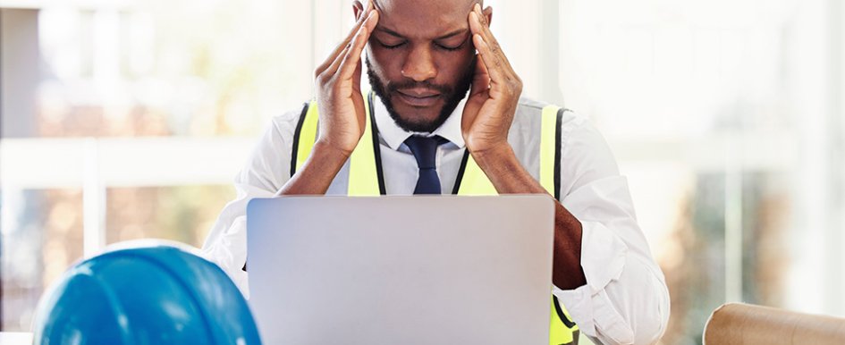 Man wearing vest looking at laptop and holding head in frustration with hardhat on desk