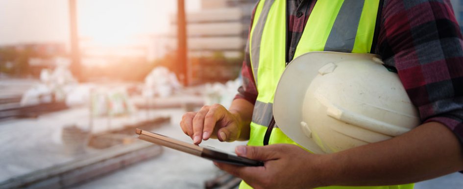 A construction worker wearing a bright safety vest has a hard hat tucked under their arm and is checking a tablet