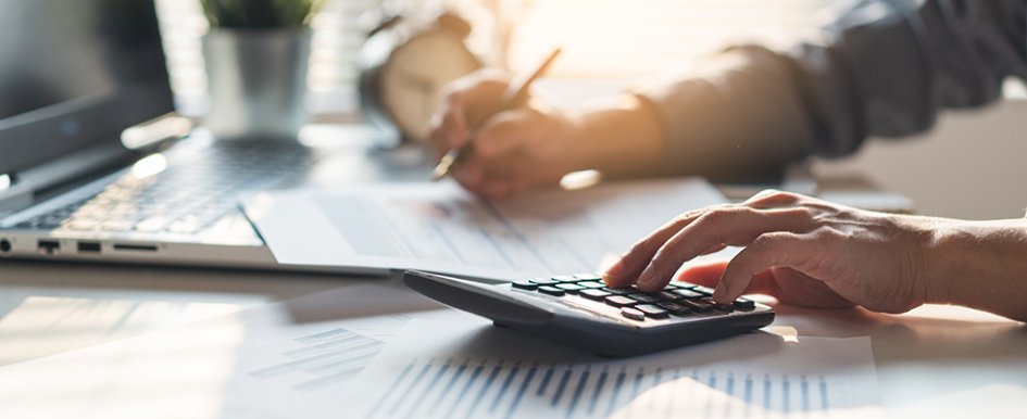 Two hands on a desk; one hand holding pencil, the other using calculator