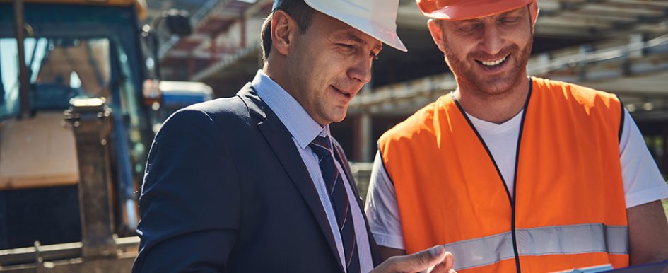 Man in orange vest and hard hat talks to man in suit and hard hat