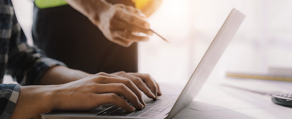 Hands on laptop while a person with hard hat and pencil gestures toward screen