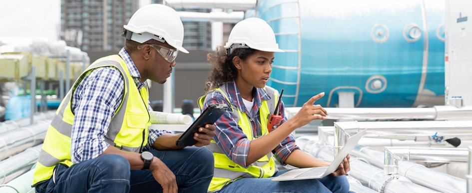 Two workers in vests and hard hats. Both looking at computer and talking