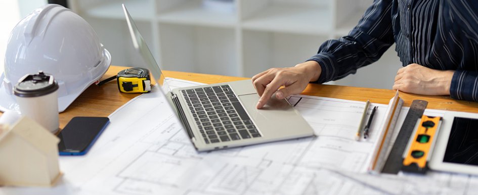 Hands using laptop on desk with hard hat, tablet, measuring tape and tools