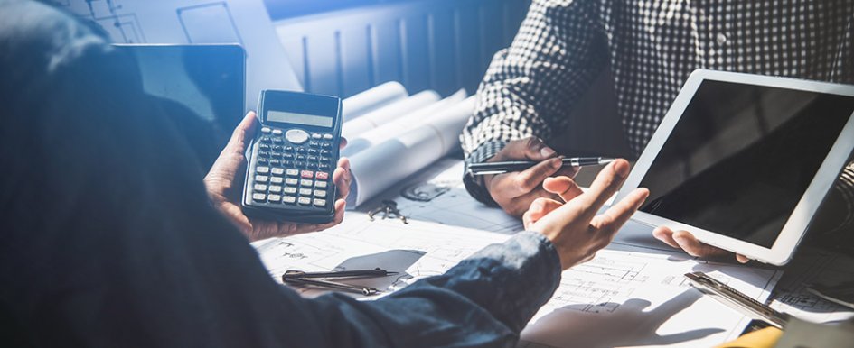 Two people sitting at desk one; one holds calculator, the other holds a pen and tablet