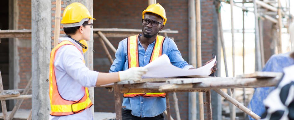 Two people wearing hard hats and bright safety vests talk over a large sheet over paper on a construction site