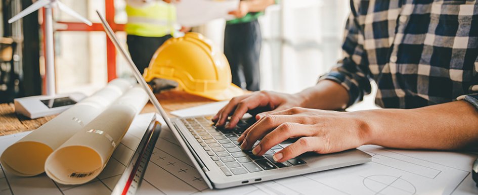 hands typing on laptop with hard hat in background