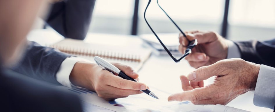 Two sets of hands point to graphs on a sheet of paper. One hand holds a pen. One hand holds a pair of glasses.