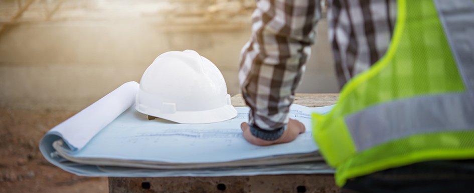 Worker in green vest working on paper plans with white hardhat in background