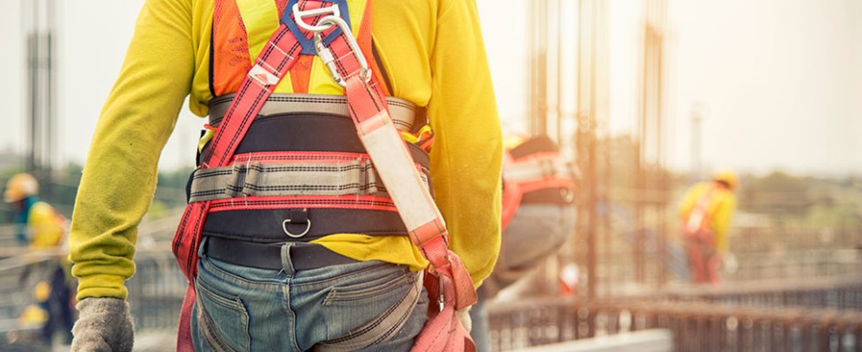 A construction worker is walking away from the camera, wearing a safety harness