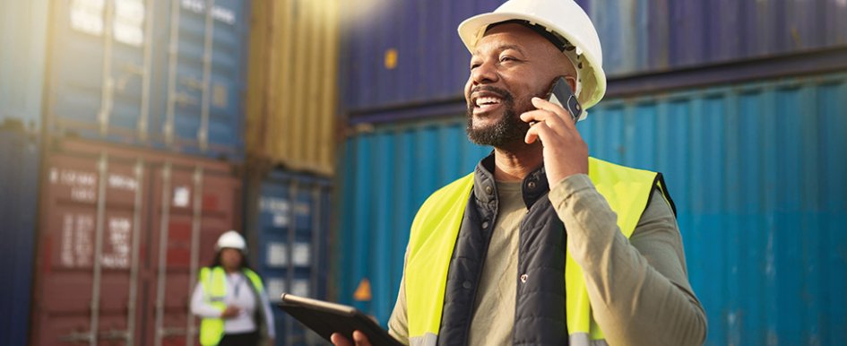 Worker in hard hat and vest using phone & smiling/Adobe Stock