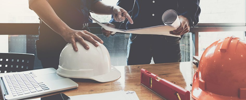 Two workers in office looking at plans; hard hats on desk 