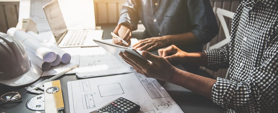 Two people sitting at desk, looking at the same tablet. Hard hat and blueprints on desk 