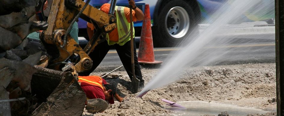 Water spews out of ground while two workers inspect nearby
