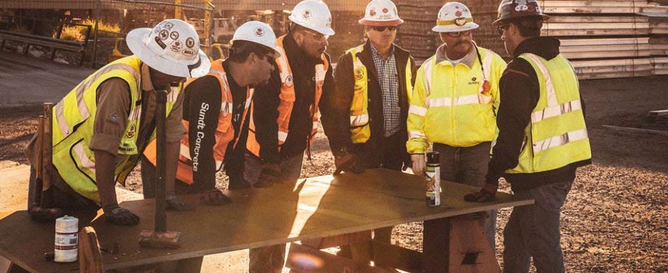 Six men in construction uniforms stand around a table