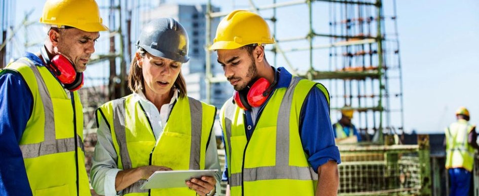 Three individuals in hard hats and bright safety vests look at a tablet display while standing on a construction site