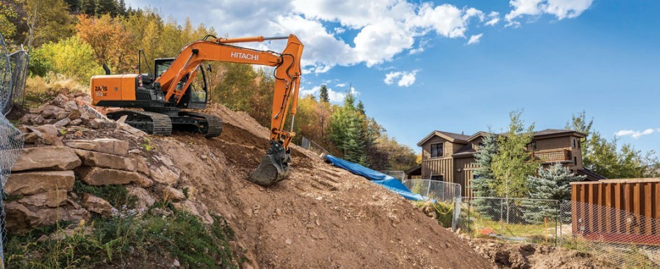 An excavator digs on the side of a hill with a house in the background