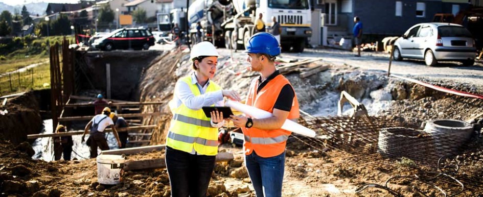 Two people in hard hats and bright safety vests are having a conversation while standing on a construction site