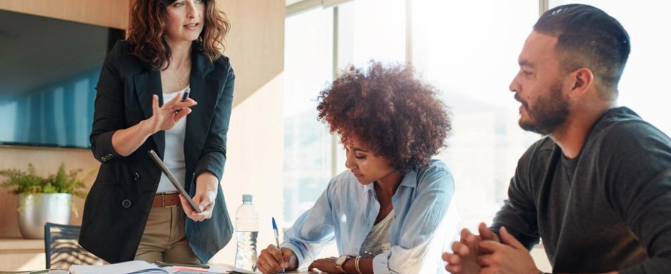 A man and a woman sit at a table, while a woman stands and shows them something on a tablet. The woman sitting is taking notes.