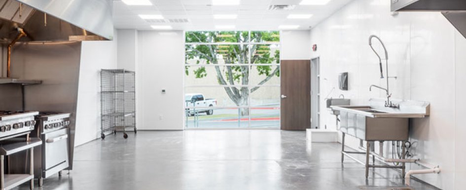 A bright room features an industrial work sink and two stovetops and ovens. A set of wire shelves sits in the back corner. A large window is at the back of the room near an open door. The floor is gray concrete and the walls are white.