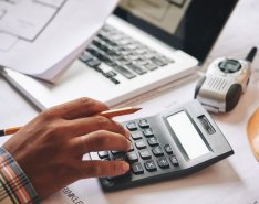 A person's hand is keying in numbers on a calculator. The hand is also holding a pencil. A portable radio and laptop can also be seen in the background.