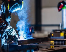 A worker welds something, illuminated in blue light