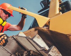 Worker in orange vest and hard hat with yellow headset, working on yellow piece of construction equipment