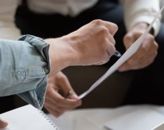 A hand uses a pen to point to a piece of paper that another set of hands is holding