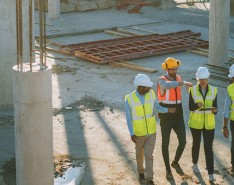 Four individuals are on a construction site. One holds a tablet; two are pointing. All four wear hardhats and bright safety vests.