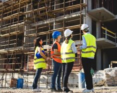 Four individuals look up at a building under construction. Three wear hardhats, but one does not.